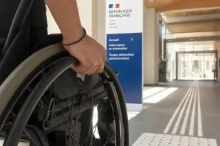 Detail of a wheelchair user's hand on the wheel beside tactile paving in a bright, accessible public building entrance.