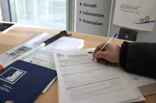 Close-up of a hand completing paperwork in a softly lit French office, with a white cane and Braille signage on the desk, evoking autonomous accessibility during administrative procedures.