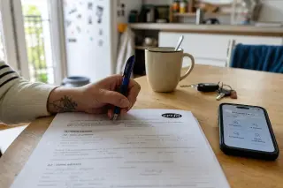 Hand annotating a French administrative document at a kitchen table with a smartphone showing an accessibility feature, soft daylight and everyday objects in the background.