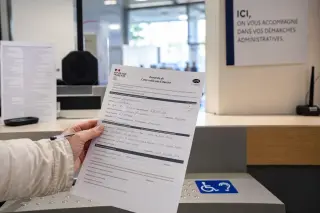 Hand holding a filled CERFA form and pen on an administrative office counter with tactile markers, neutral light and signage in a modern French setting.