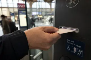 A natural close-up of a hand inserting a mobility card with braille into a public transportation ticket machine, captured in a vibrant but calm French train station.