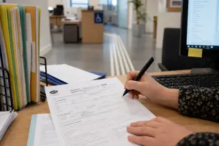 Close-up of hands filling out a CERFA form in a public office, with a tactile guidance path visible in the background