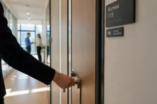 Hand opening an office door with accessible signage in a sunlit modern workplace corridor, subtle interaction between colleagues in the background.