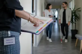 Close-up of a hand holding a binder with a 'Référent handicap' badge in an office corridor with two colleagues in the background, one using a white cane, in soft natural daylight.