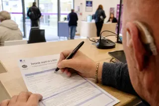 Close-up of a hand annotating a CERFA form at a French administrative counter, with a discreet hearing aid and digital device visible, soft natural light, blurred agency background